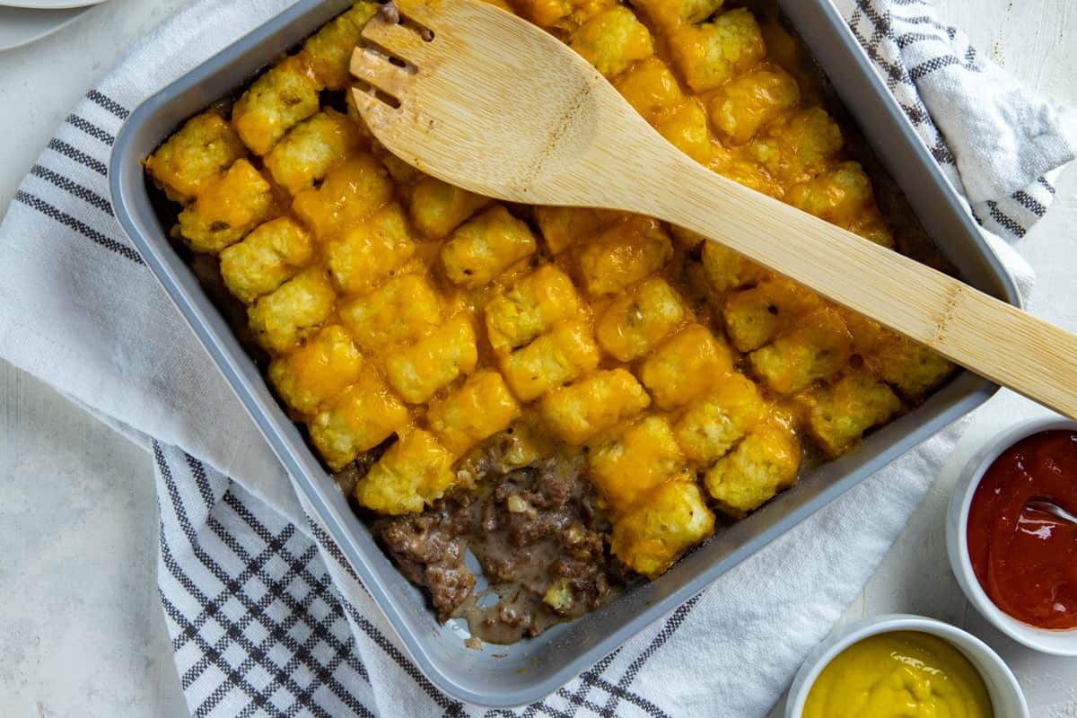 Baked tater tot casserole in square metal pan with scoop removed from corner. Mustard and ketchup in small round white bowls on the side.  Wooden serving spoon resting on top of casserole.