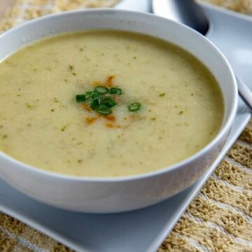 Soup in round white bowl sitting on white square plate. Soup topped with sliced green onions.