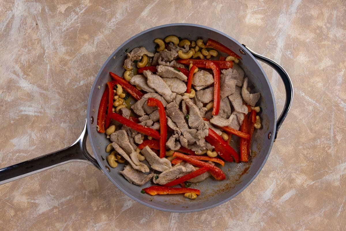 Sliced pork, bell pepper strips, and cashew nuts that have been sauteed in a large skillet.
