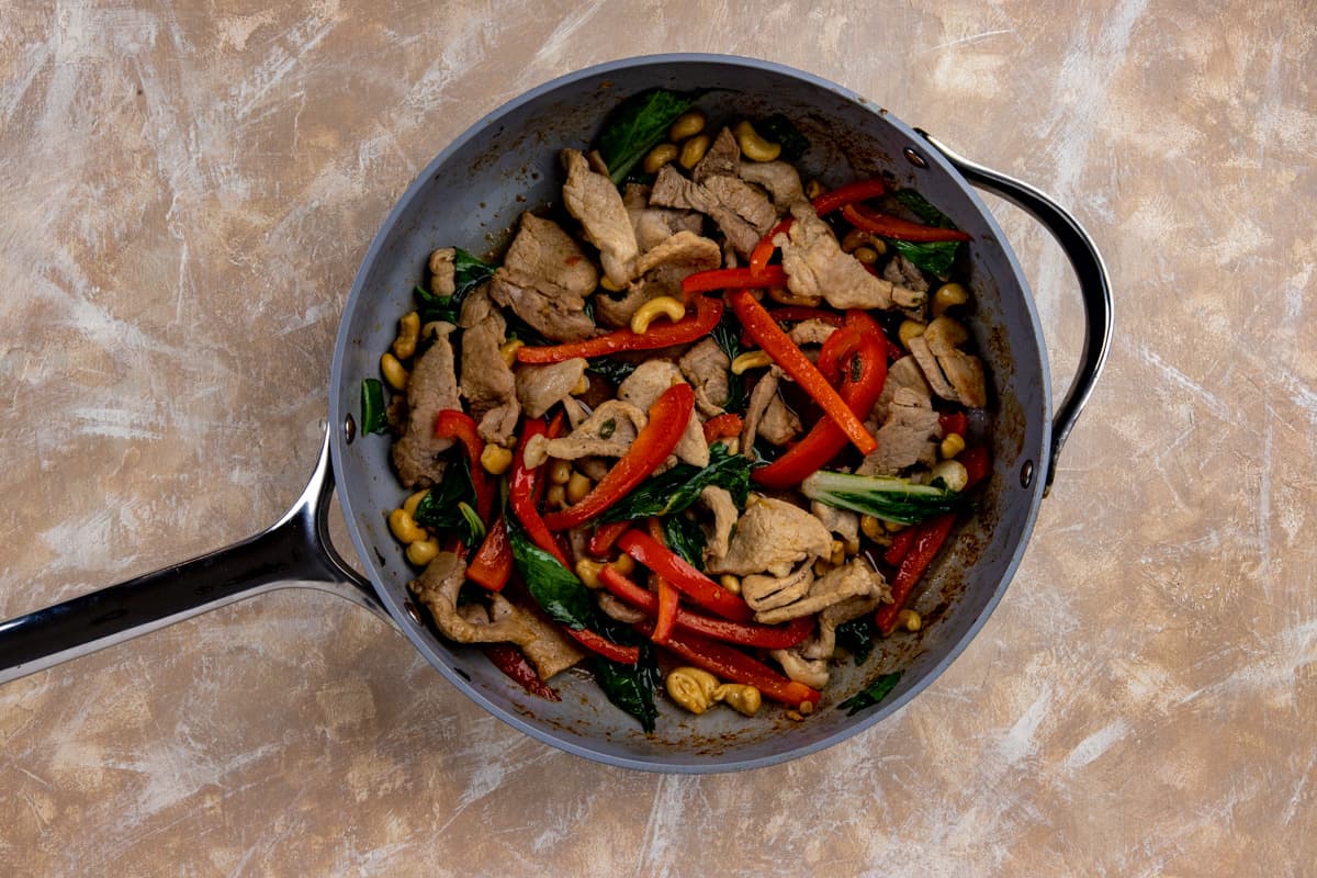 Sliced pork, bell pepper strips, cashew nuts, and seasonings that have been sauteed in a large skillet.
