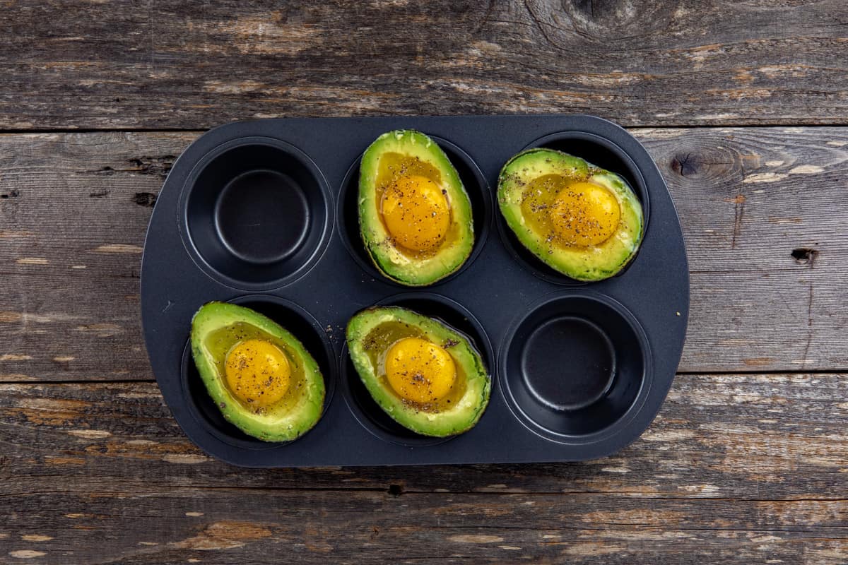 Four avocado halves with a raw egg, salt, and pepper sprinkled on top in each muffin cup in a muffin pan.