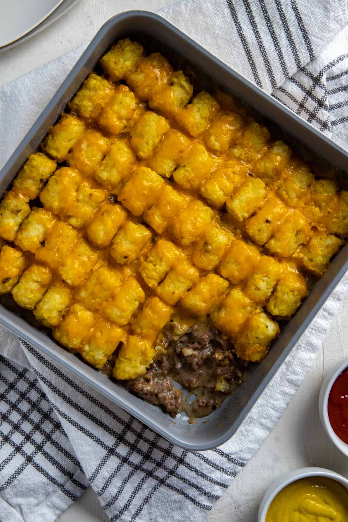 Baked tater tot casserole in square metal pan with scoop removed from corner.  Mustard and ketchup in small round white bowls on the side.