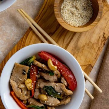 Finished stir fry in white bowl with wooden chop sticks and small bowl of toasted sesame seeds on the side.