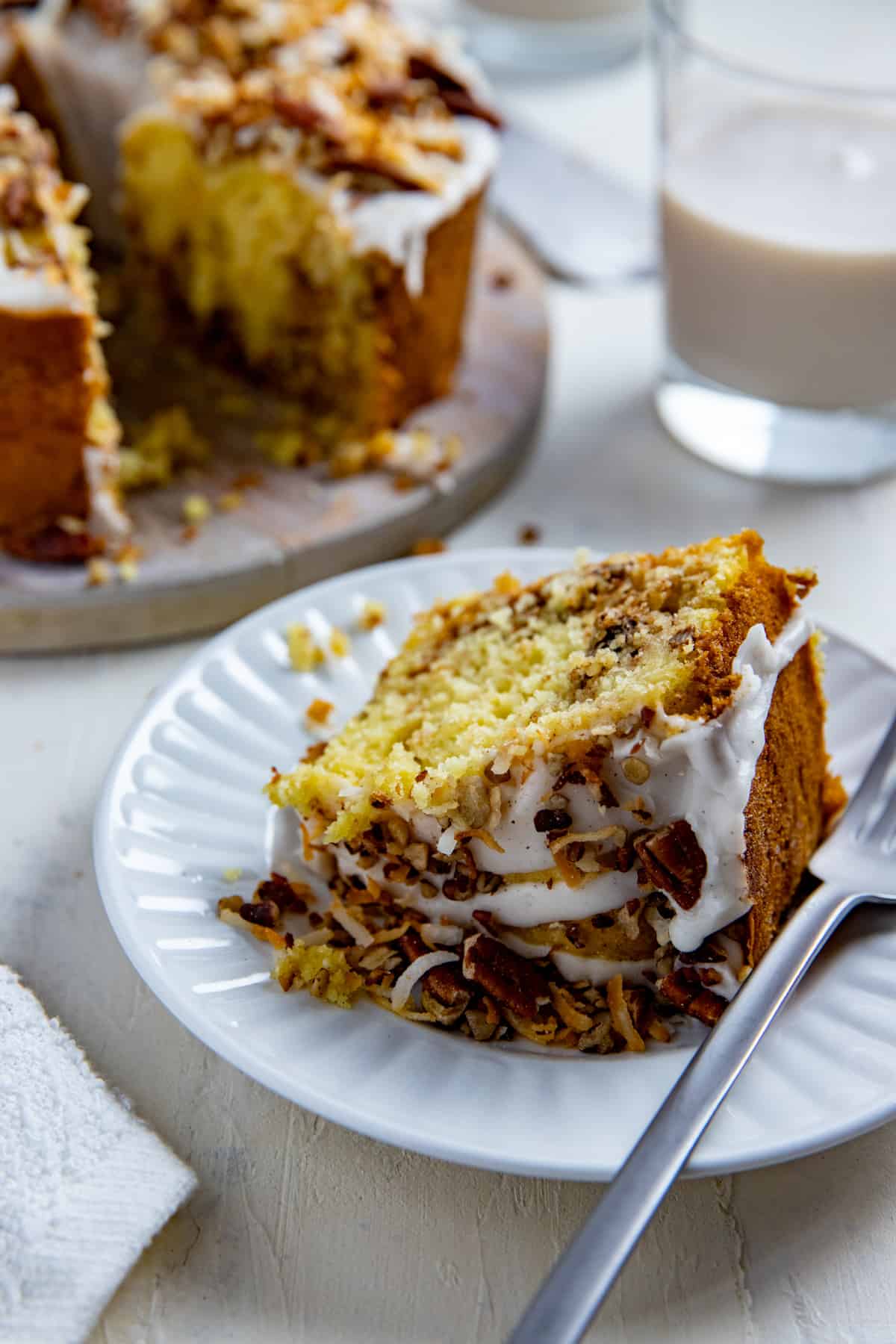 Slice of coconut coffee cake on white plate with fork.  Whole cake in background with glass of milk.