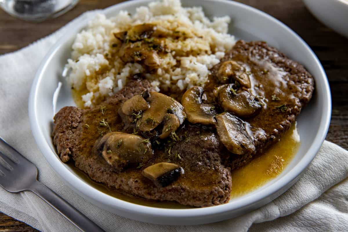 Two cooked steaks covered with gravy and mushrooms with a side of rice and gravy.