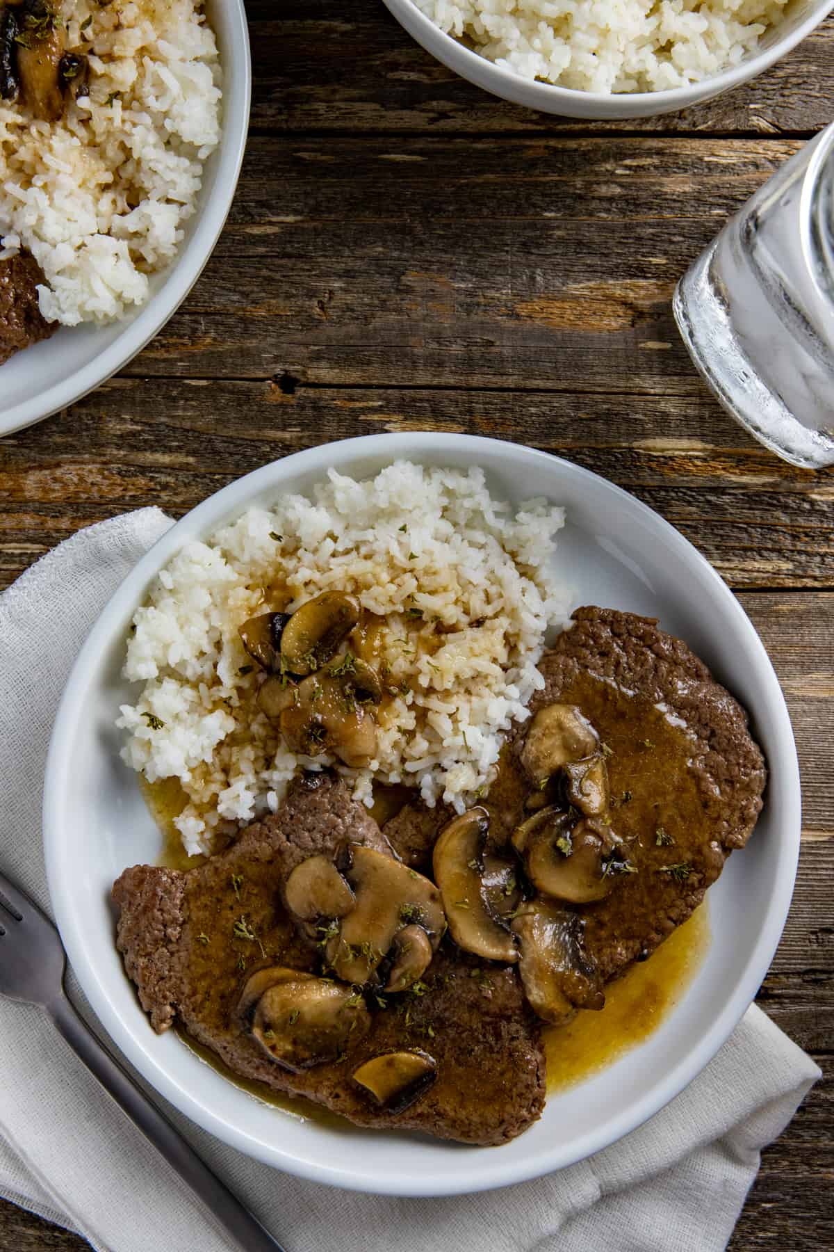 Two cooked steaks covered with gravy and mushrooms with a side of rice and gravy.