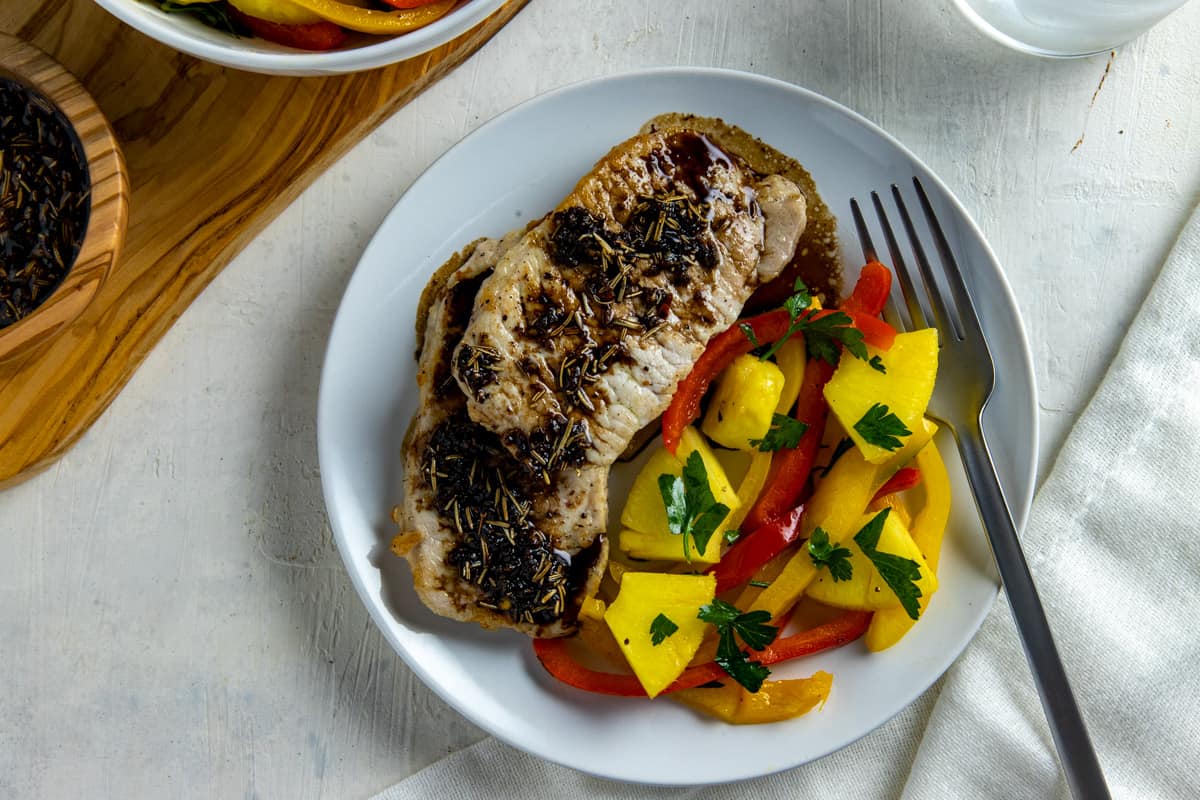 Two cooked pork chops topped on a white plate with glaze and pineapple chunks with sliced bell peppers on the side.