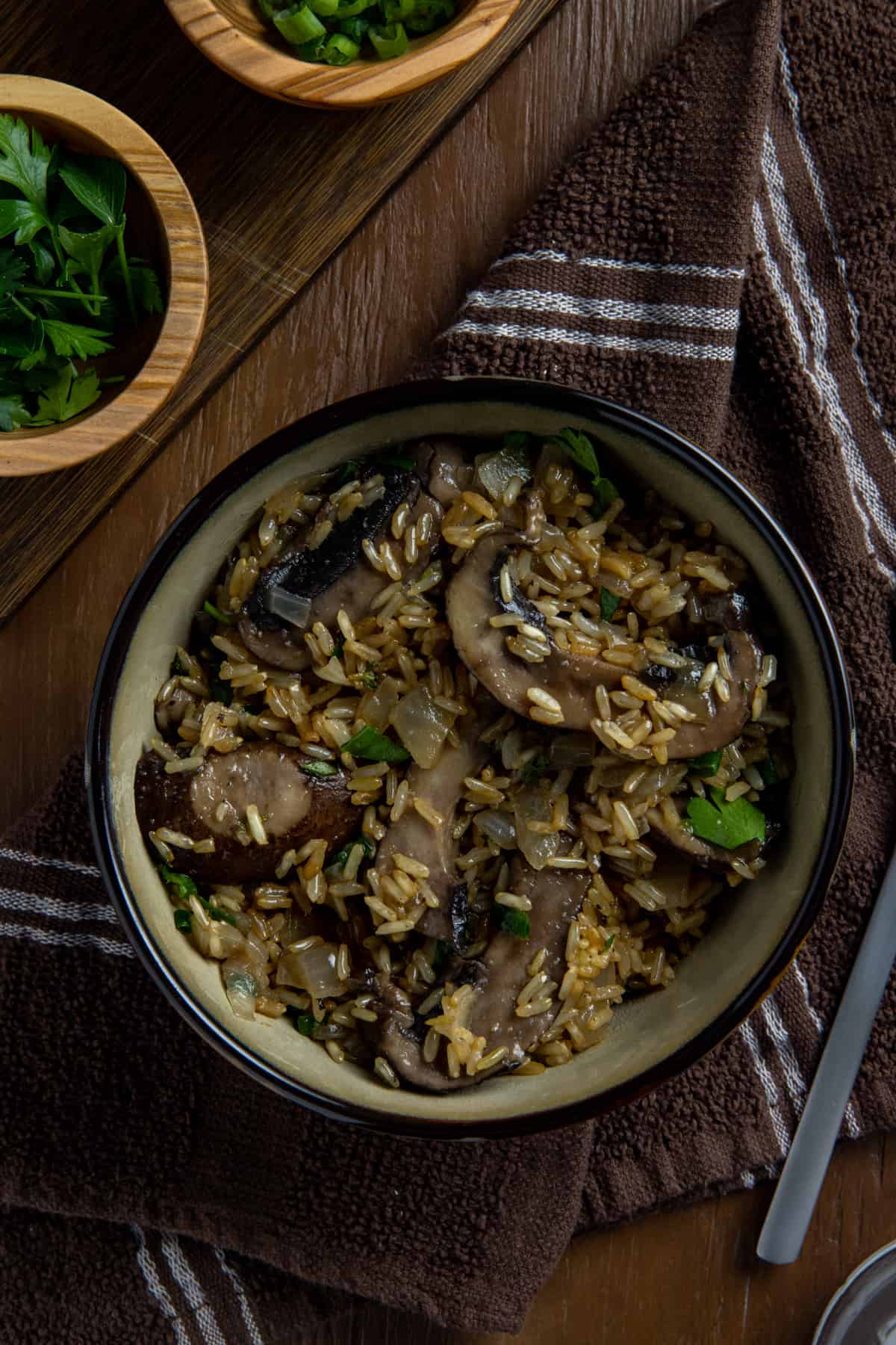 Cooked mushroom rice in brown bowl with spoon on the side.  Small bowl of sliced green onion in the background.