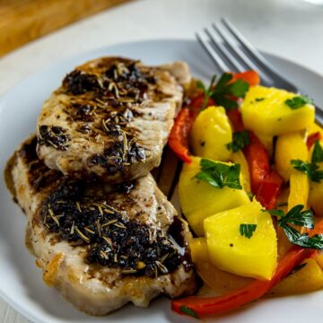 Two cooked pork chops topped on a white plate with glaze and pineapple chunks with sliced bell peppers on the side.