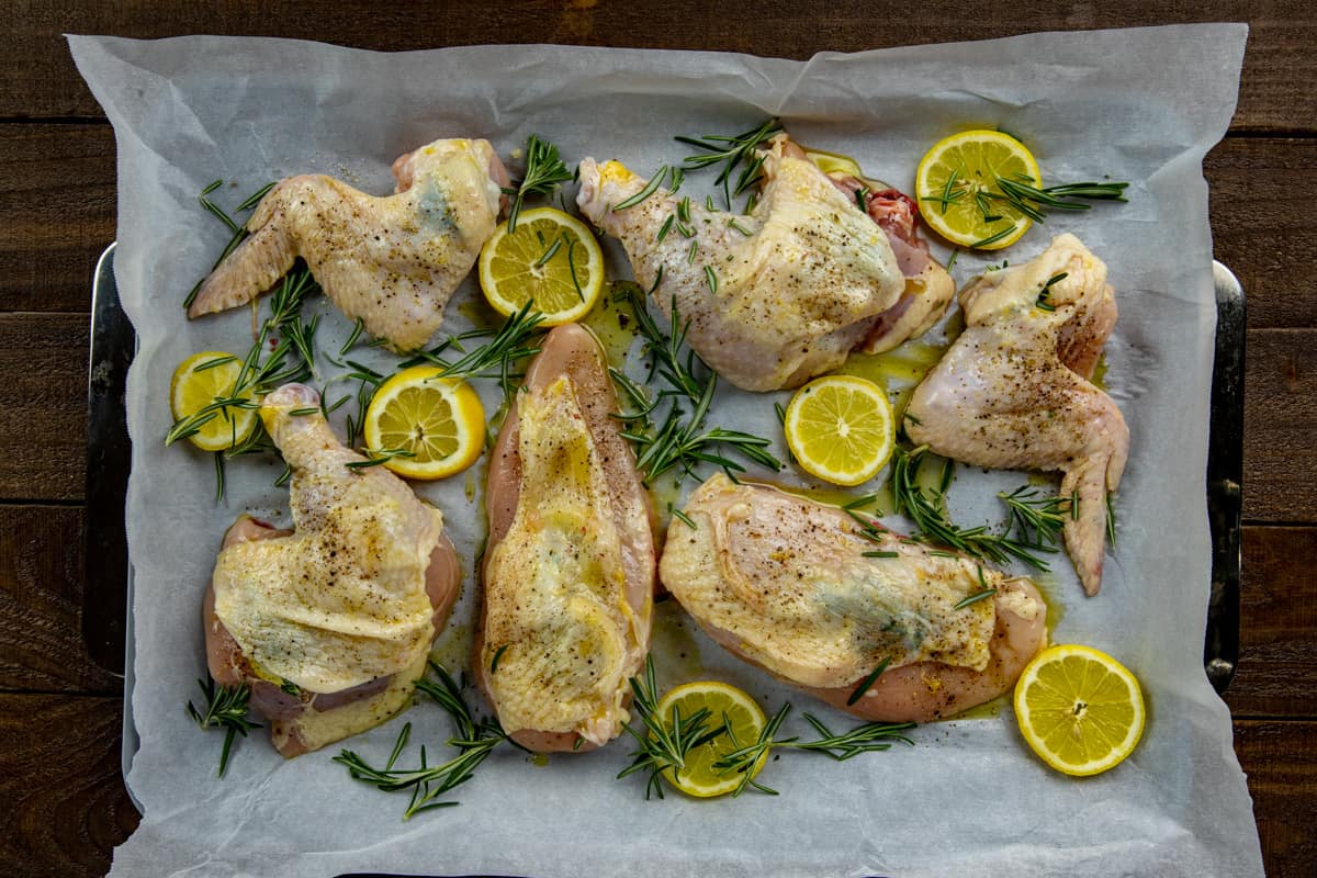 Uncooked chicken pieces topped with lemon slices and rosemary sprigs on a sheet pan.