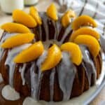 Finished cake topped with peaches and glaze in a rimmed cake platter with glaze running down the cake and into the platter.