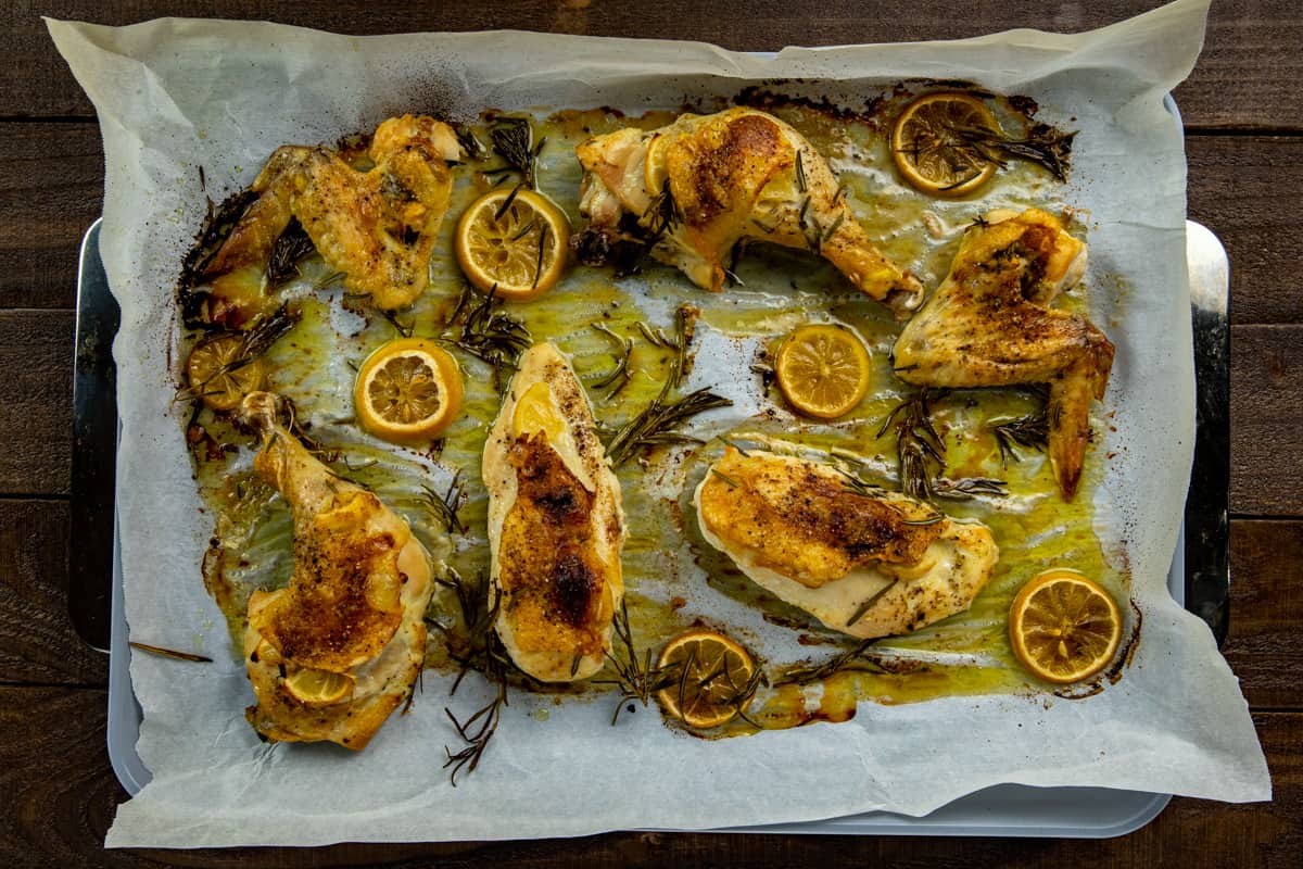 Roasted chicken pieces topped with lemon slices and rosemary sprigs on a sheet pan.