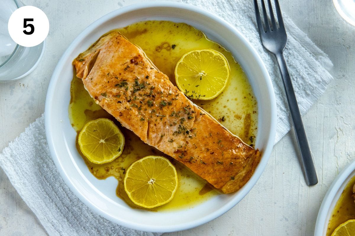 Cooked salmon fillet on a white plate with lemon butter sauce and 3 lemon slices.