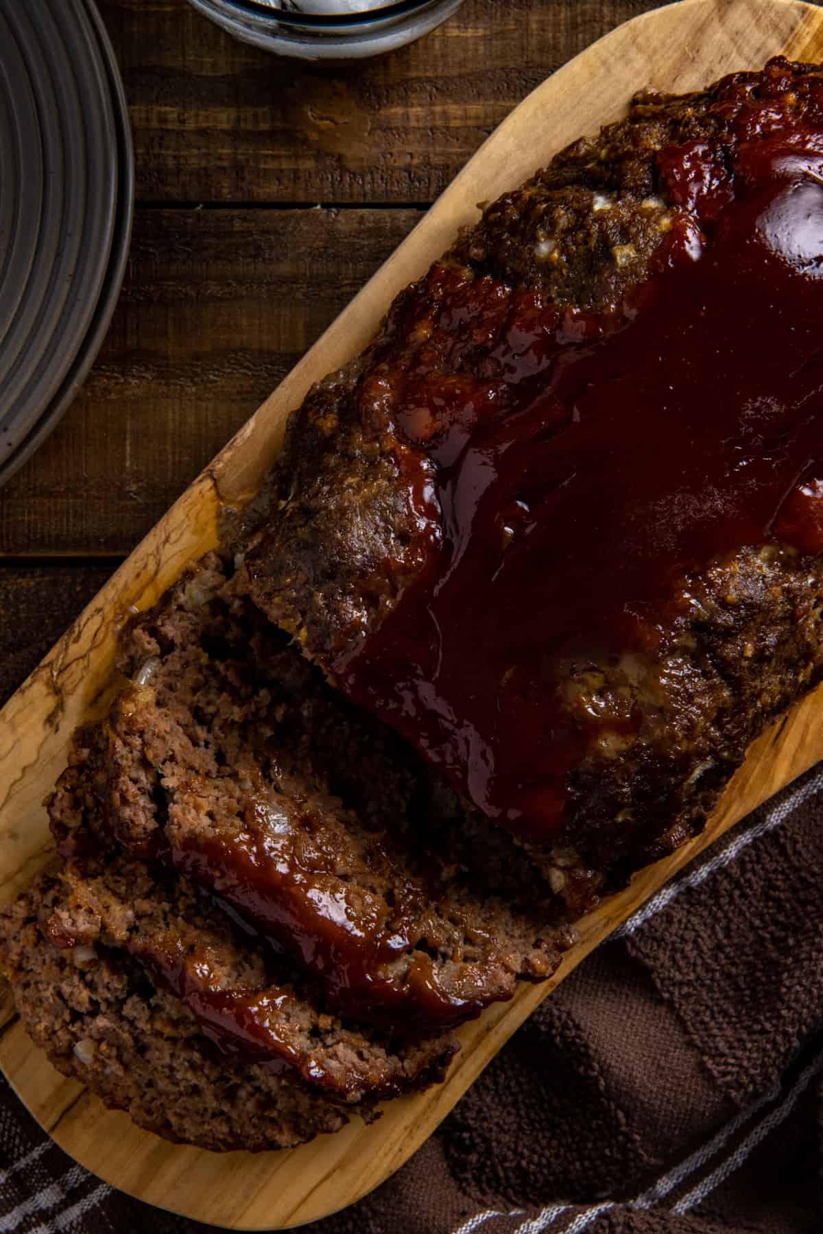 Baked meatloaf on wooden platter with a few slices revealed. Meatloaf sauce on top and running down the sides a little.