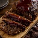 Baked meatloaf on wooden platter with a few slices revealed. Meatloaf sauce on top and running down the sides a little.