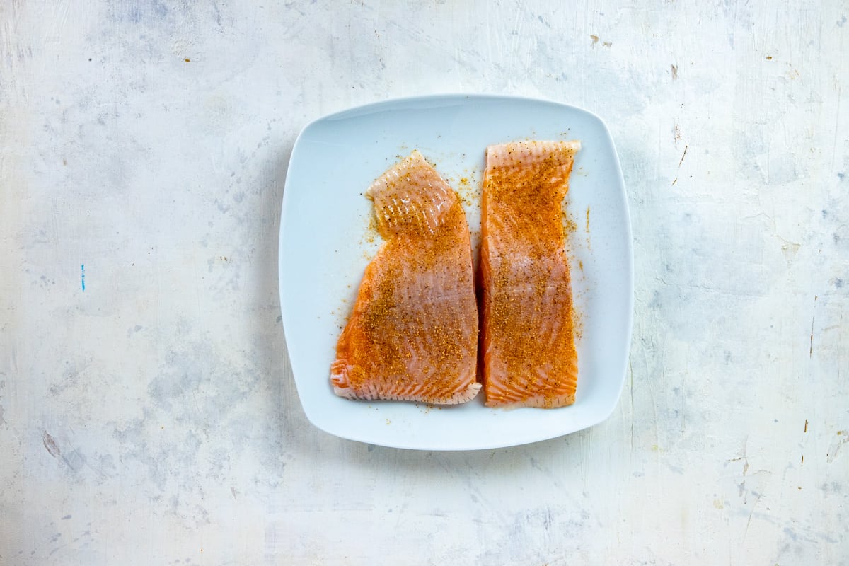 Two raw salmon fillets sprinkled with Cajun seasoning on white plate.