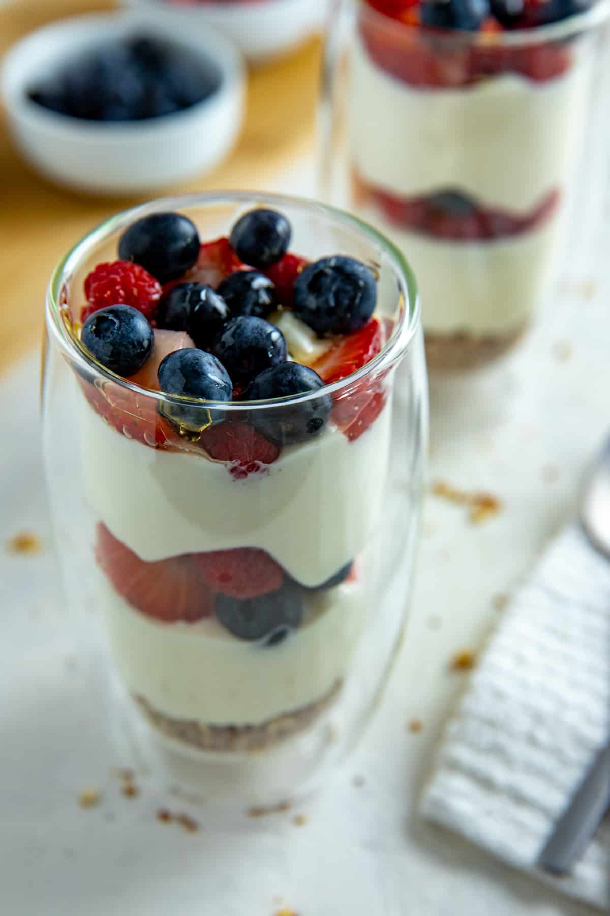 Two tall clear glasses with a granola crust at the bottom, two layers of yogurt and two layers of strawberries and blueberries.