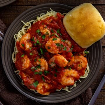 Shrimp Marinara spaghetti on a brown plate with a dinner roll on the side.