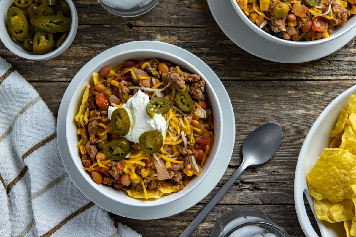 Taco soup in white bowl garnished with corn chips, sour cream, and jalapeno slices.