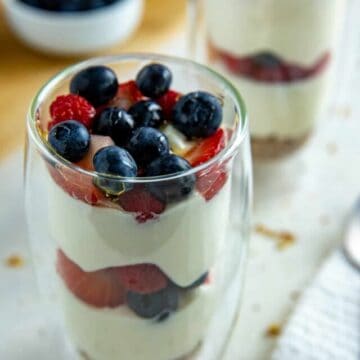 Two tall clear glasses with a granola crust at the bottom, two layers of yogurt and two layers of strawberries and blueberries.