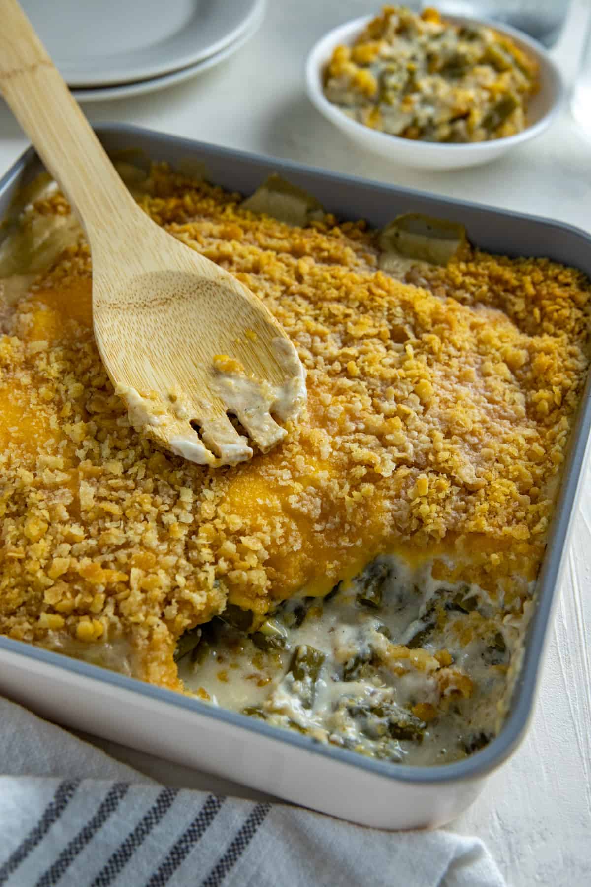 Cooked green bean casserole in square casserole dish with one serving removed.  A wooden serving spoon is resting on top of the casserole.