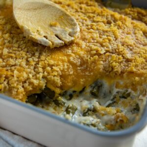 Cooked green bean casserole in square casserole dish with one serving removed. A wooden serving spoon is resting on top of the casserole.