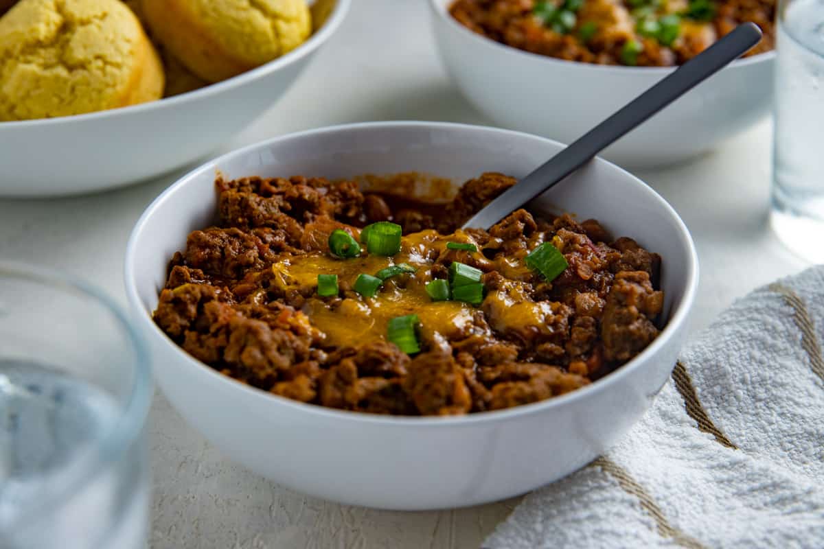 Cooked chili in white bowl topped with shredded cheddar cheese and green onion slices.