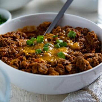 Overhead view of cooked chili in a white bowl topped with cheese and sliced green onions. Cornbread muffins in the background.