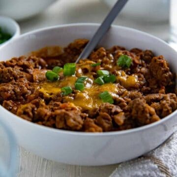 Cooked chili in white bowl topped with shredded cheddar cheese and green onion slices.
