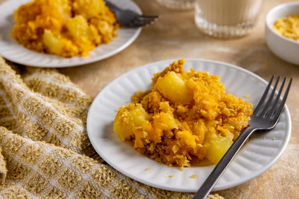 A white plate holds a serving of pineapple casserole topped with a golden, crumbly crust. A fork rests on the plate. Another plate with the same dish and a checked cloth are visible in the background. The setup is on a beige surface.
