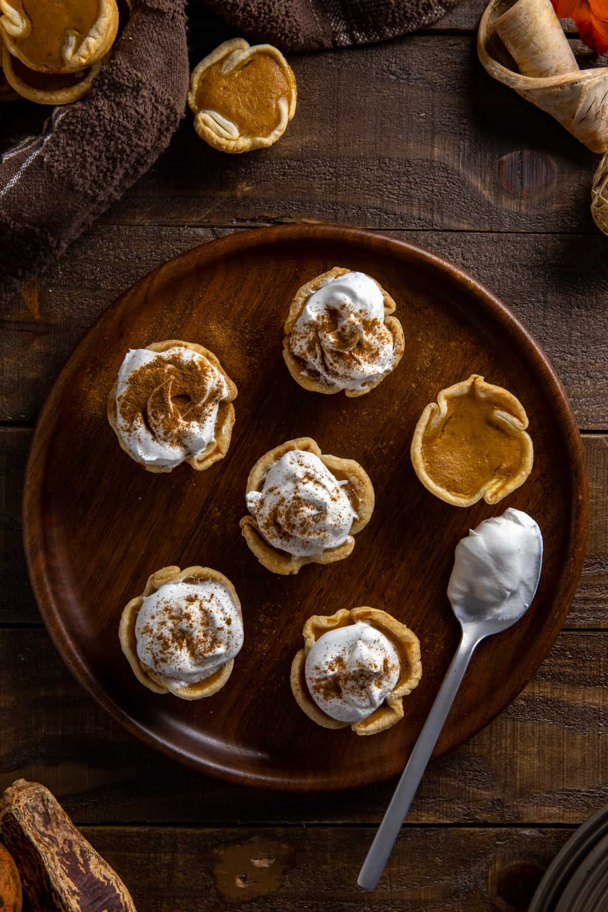 Six baked mini pumpkin pies on round wooden plate with spoonful of whipped topping on the side.