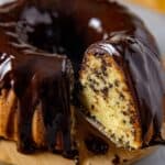 Baked chocolate chip pound cake with chocolate ganache on round wooden cake platter. One slice being removed with a spatula.