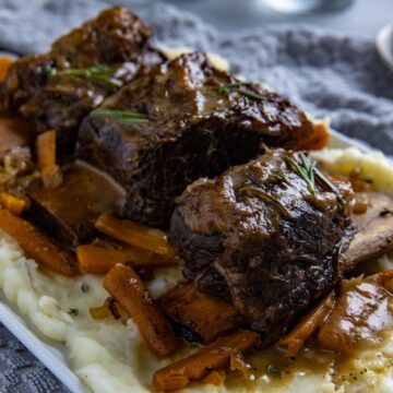 Cooked beef ribs on a bed of mashed potatoes with onions and carrots scattered around.