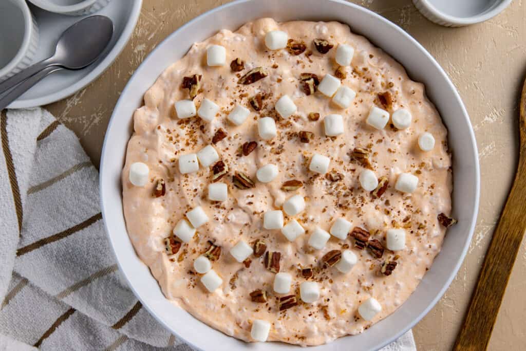 A bowl of peach fluff salad is topped with mini marshmallows and pecan pieces. The dessert is creamy and light orange in color. The bowl sits on a beige surface, accompanied by a striped towel, two spoons, and a small white dish.