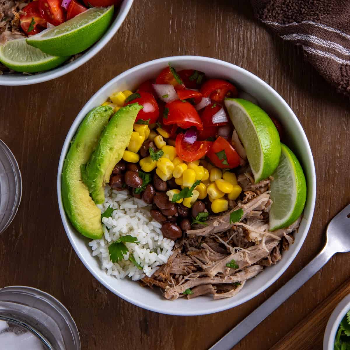 White bowl filled with pork carnitas, black beans, corn, salsa, garnished with lime slices and avocado wedges.