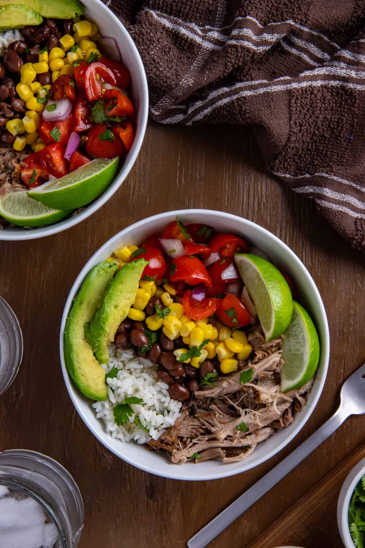 Two white bowl filled with pork carnitas, black beans, corn, salsa, garnished with lime slices and avocado wedges.