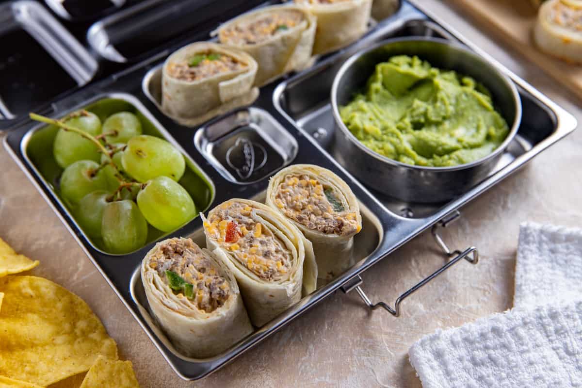 Taco meat pinwheels in a stainless steel meal prep container with guacamole and green grapes on the side.