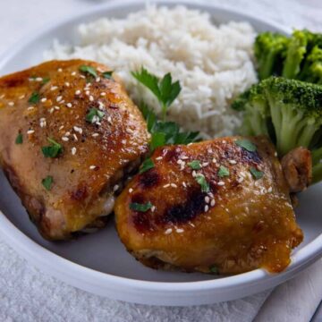 Two baked chicken thighs on a white plate with cooked white rice and steamed broccoli on the side.