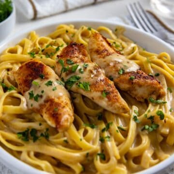 Blackened Chicken Alfredo in a white bowl. Fettucine tossed in alfredo sauce with 3 blackened chicken tenderloins on top. Alfredo sauce drizzled on top of the chicken and fresh parsley sprinkled on top.