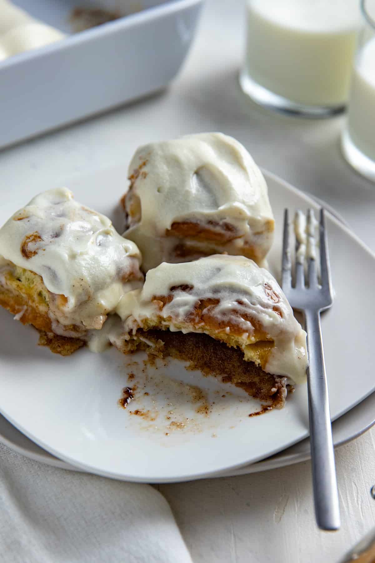Three Hawaiian Roll Cinnamon Rolls on a square white plate with a fork on the side and a glass of milk in the background.