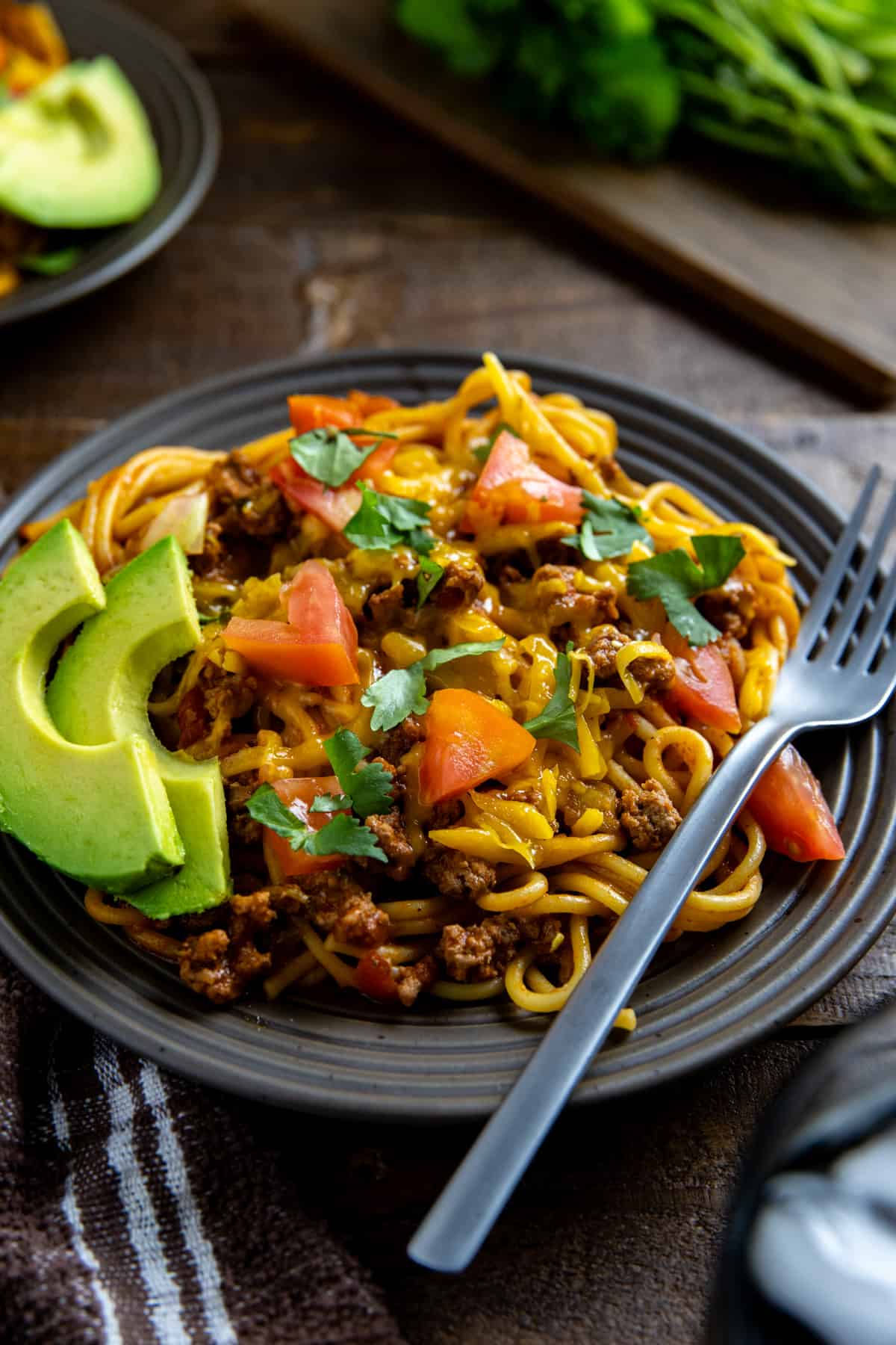Mexican spaghetti topped with chopped tomatoes and fresh cilantro on brown plate with fork on the side.  Avocado slices on the side.