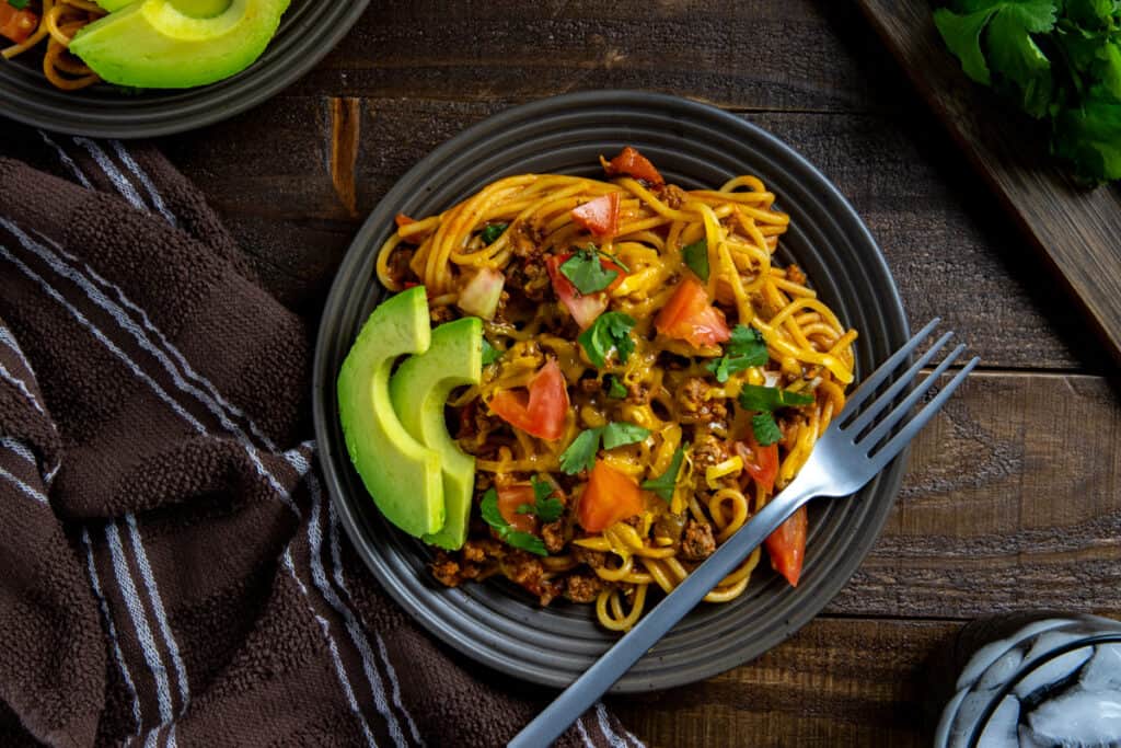 A plate of spaghetti topped with ground beef, diced tomatoes, and herbs. Sliced avocado is placed on the side. A fork rests on the plate, which is set on a wooden table with a brown striped cloth nearby.