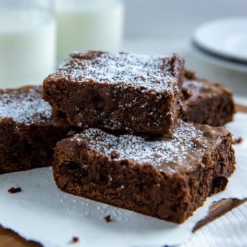 Four baked brownies sprinkled with powdered sugar on a wooden platter with two glasses of milk in the background.