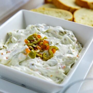 Olive dip in a square, white serving bowl topped with chopped green olives. Crostini in the background.