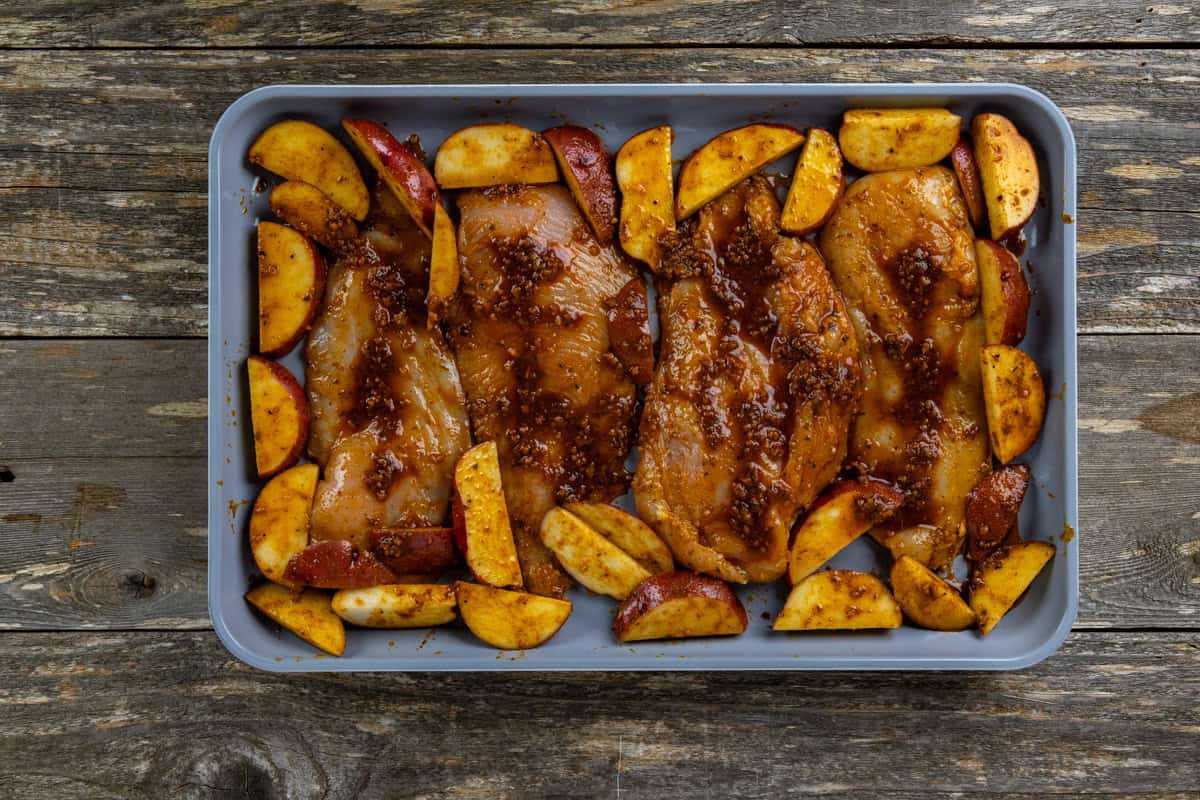 Uncooked seasoned chicken and potato wedges on a sheet pan.