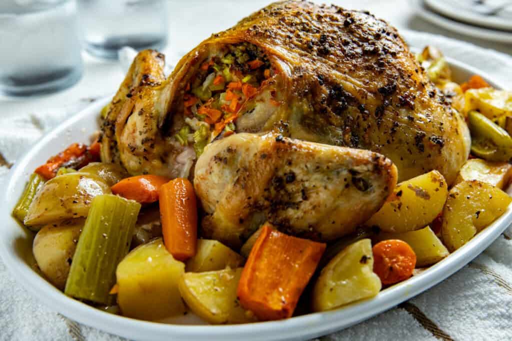 A roasted stuffed chicken on a platter, surrounded by roasted vegetables including potatoes, carrots, celery, and onions. The chicken is golden brown with visible herbs and spices. A couple of drinking glasses are in the background.