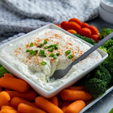 Prepared dip in a white square dish on a white platter with carrots sticks, celery sticks, broccoli florets, and cherry tomatoes.