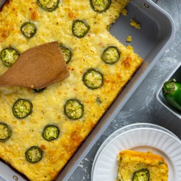 Baked casserole in a baking dish with one serving removed. One serving on a white plate. Wooden spoon resting on top of the casserole.