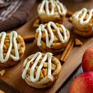 Five baked and frosted apple pie cinnamon rolls on wooden cutting board with some cinnamon sticks and fresh red apples on the side as garnish.