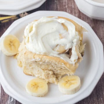 One frosted cinnamon roll on a white plate with some fresh sliced banana on the side.